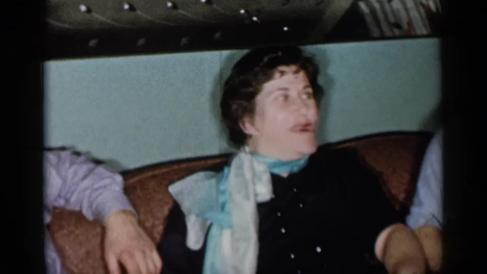 A woman wearing a pale scarf over a dark top sits in a 1955 New York City living-room frame, looking upward during conversation.