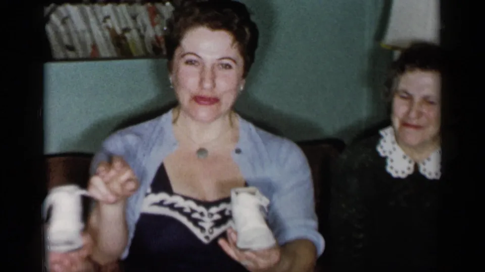 1955 New York City living-room frame showing a woman presenting small white baby shoes while another woman smiles nearby.