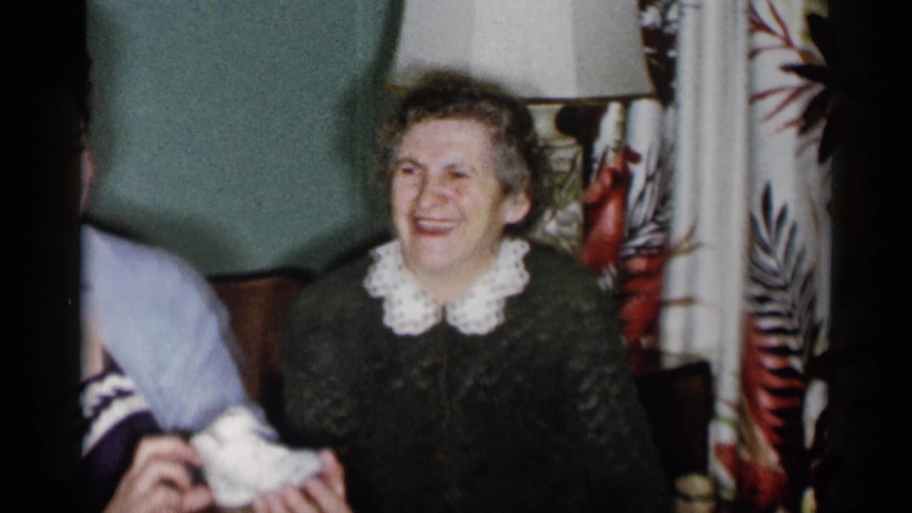 Older woman smiling in a living room beside a partly visible younger woman holding baby shoes.