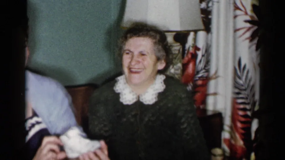 In a 1955 New York City home-movie frame, an older woman in a green sweater smiles as a younger woman at the edge holds white baby shoes.