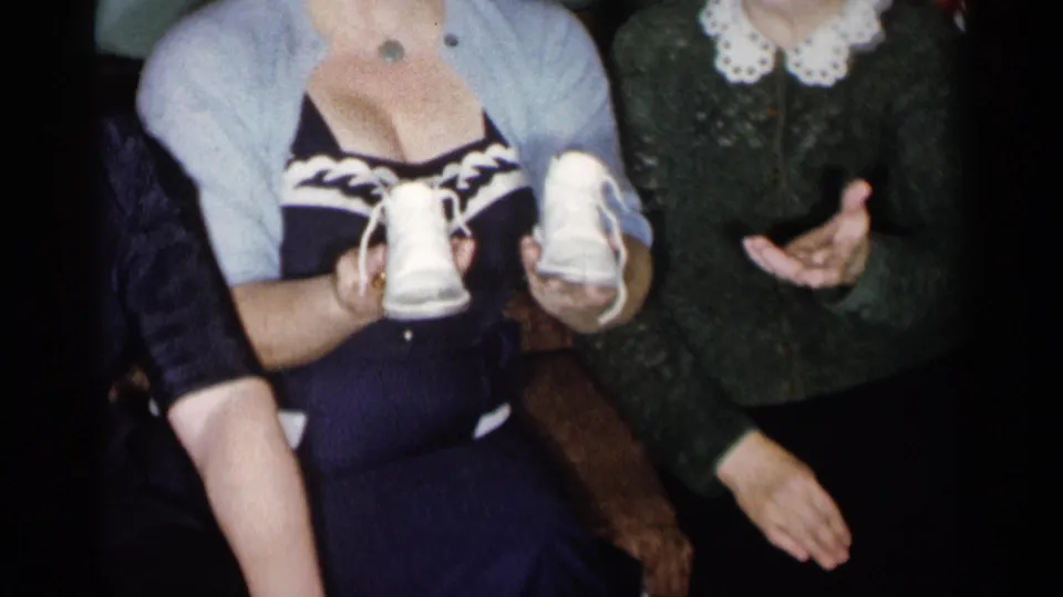 The younger woman in a blue cardigan displays tiny white baby shoes as the older woman beside her gestures in a private living room.