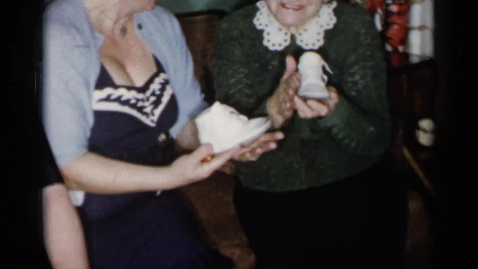 A younger woman holds out tiny white baby shoes while the older woman reaches toward them in a warmly lit 1950s living-room setting.
