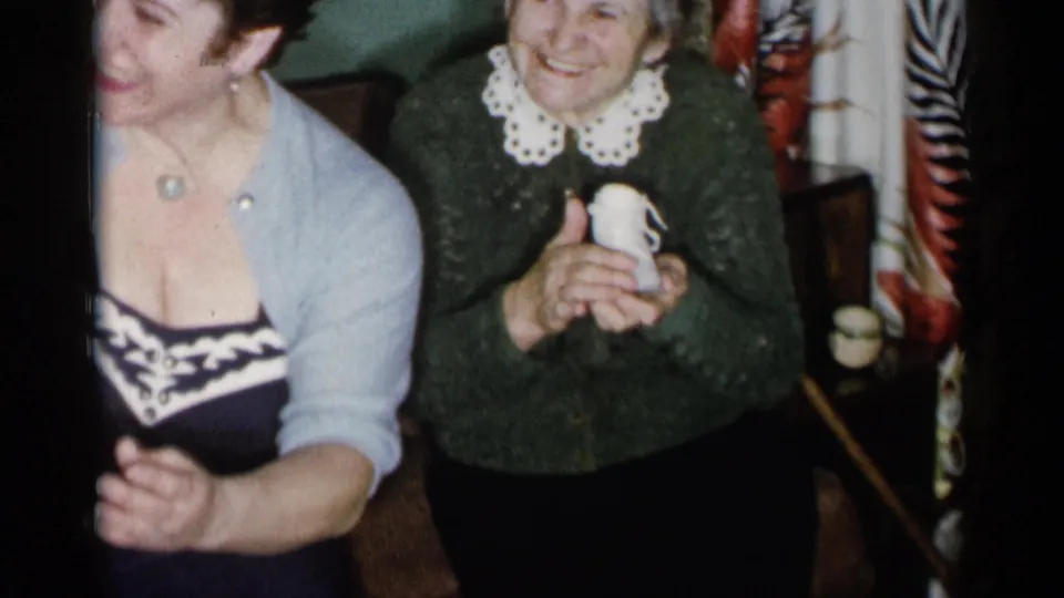 The older woman smiles and holds one tiny white baby shoe while the younger woman leans beside her in the New York City home-movie scene.