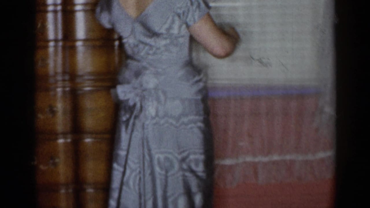 In a 1955 New York City home-movie frame, a woman in a silver-gray formal gown poses in a pink-walled room near a dresser and window.