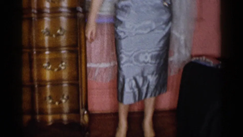 The woman is framed from shoulders to feet as she models a fitted silver-gray gown in the pink-walled room with a dresser nearby.