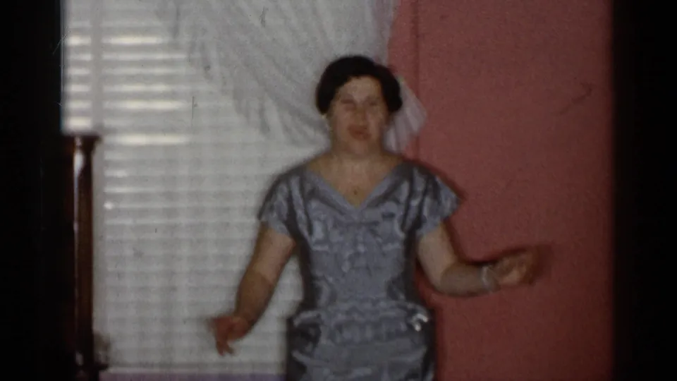 The woman faces the camera in the silver-gray formal gown, standing by white curtains and a pink wall inside the private room.