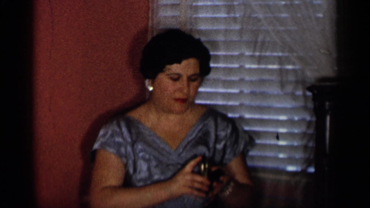 Older woman in a blue patterned dress and pearl earrings handles a small metal glass in a 1955 New York City interior.