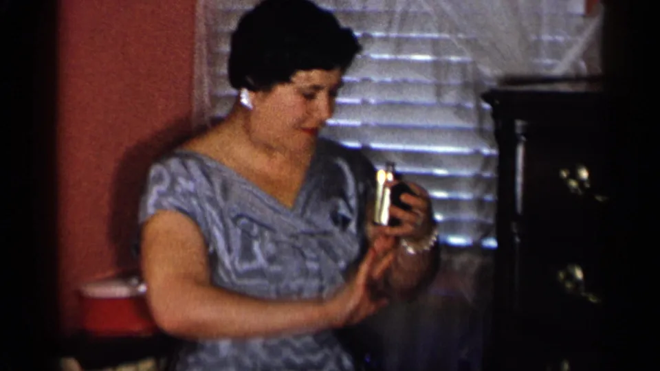In a dim 1955 New York City interior, the woman inspects or wipes a small metal glass beside blinds and a cabinet.