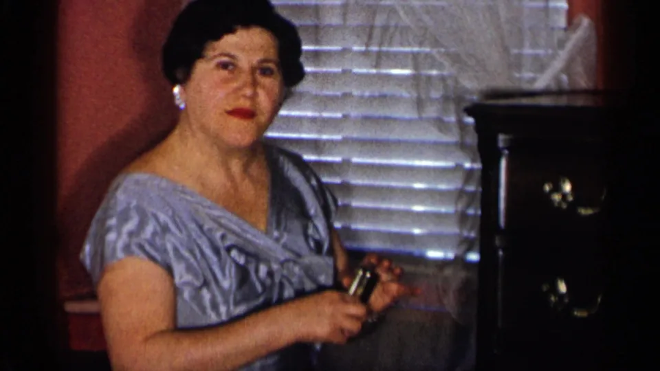 The woman briefly faces the camera while holding the small metal glass in a warm mid-century private interior.