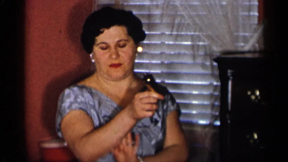 The woman continues handling the metal glass near horizontal blinds and a dark wooden cabinet in the home interior.
