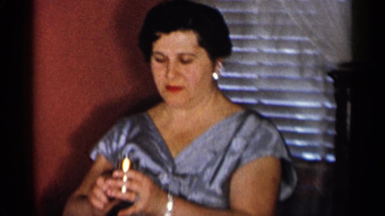 Woman in a blue dress holds lipstick near a window blind in a dim room.