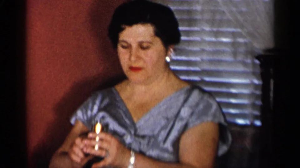 In 1955 New York City home-movie footage, a woman in a blue patterned dress holds lipstick while preparing indoors.