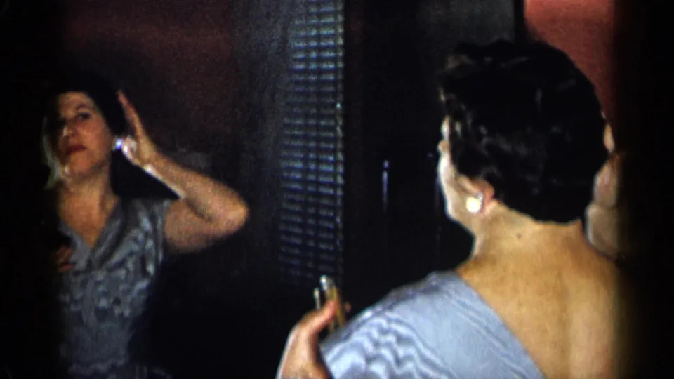 The frame shows the woman and her mirror reflection as she raises a hand to her hair during formal preparation.