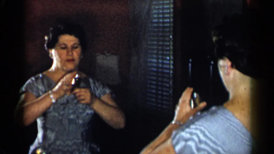 The woman faces her mirror reflection and handles a small cosmetic container, with bracelet, earrings, and blue dress visible.