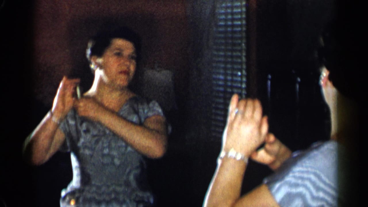 Woman in a blue patterned dress faces a mirror while touching her face and hair.