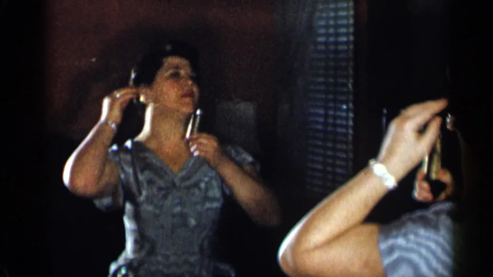 The woman stands before a mirror in a dim room, holding a small grooming item while checking her hair or makeup.