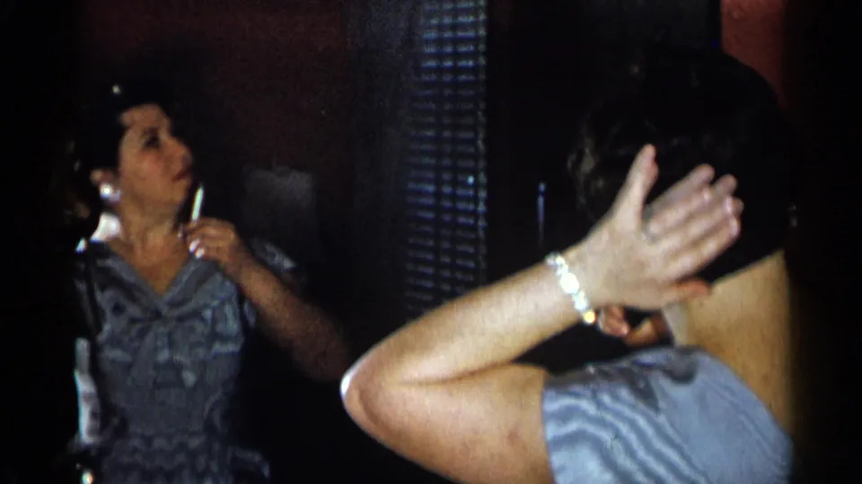 A woman in a blue patterned dress adjusts the back of her hair in front of a mirror in 1955 New York City home footage.
