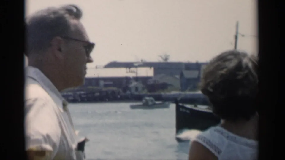A man in sunglasses and a woman seen from behind on a boat in Cape Cod, with harbor structures and water beyond.