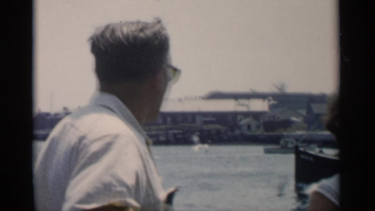 In 1956 Cape Cod home-movie footage, a man in sunglasses faces a harbor from a moving boat with docks and buildings beyond.