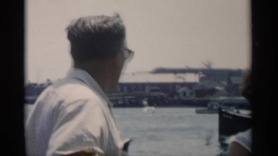 In 1956 Cape Cod home-movie footage, a man in sunglasses faces a harbor from a moving boat with docks and buildings beyond.