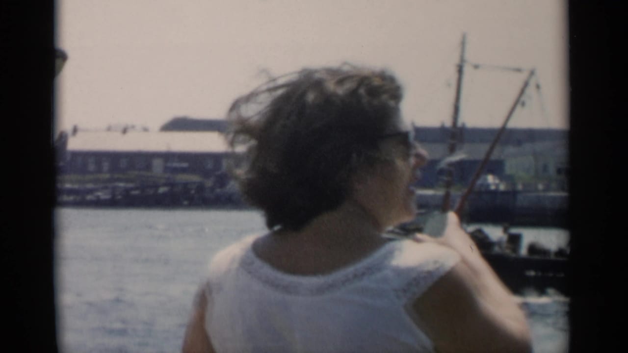 A woman in a white sleeveless top looks toward Cape Cod harbor water, with docks, a small mast, and waterfront buildings in the distance.