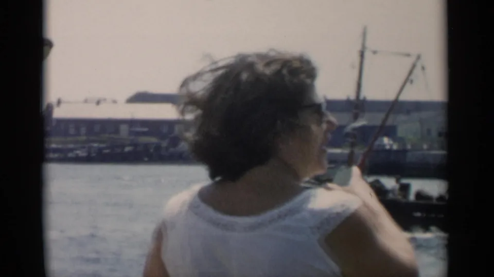 A woman in a white sleeveless top looks toward Cape Cod harbor water, with docks, a small mast, and waterfront buildings in the distance.