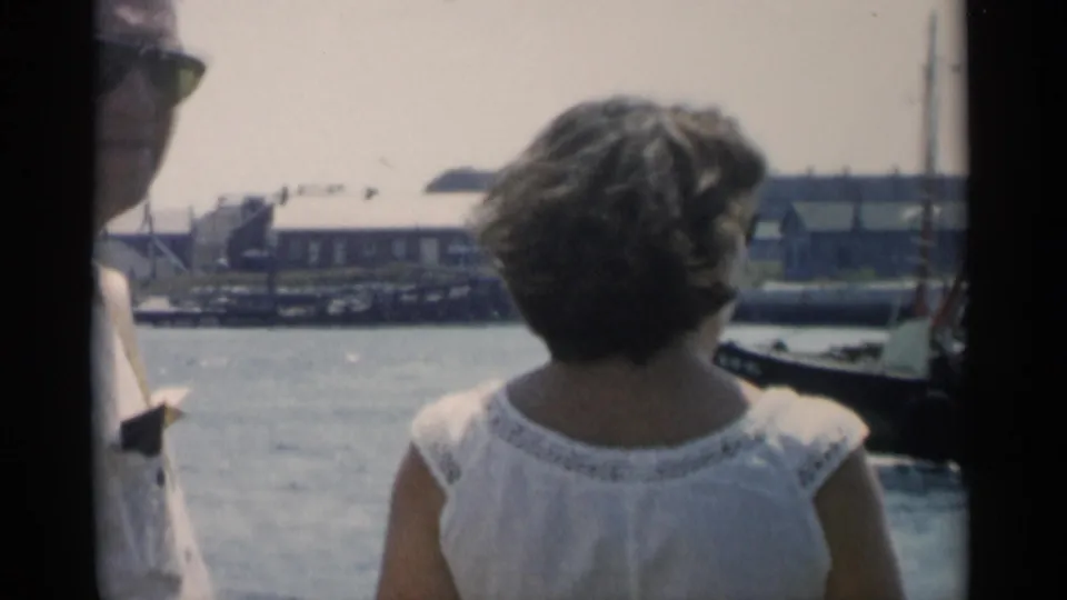 A man and woman aboard a boat look toward a Cape Cod waterfront, with calm water, docks, buildings, and dark boat framing visible.