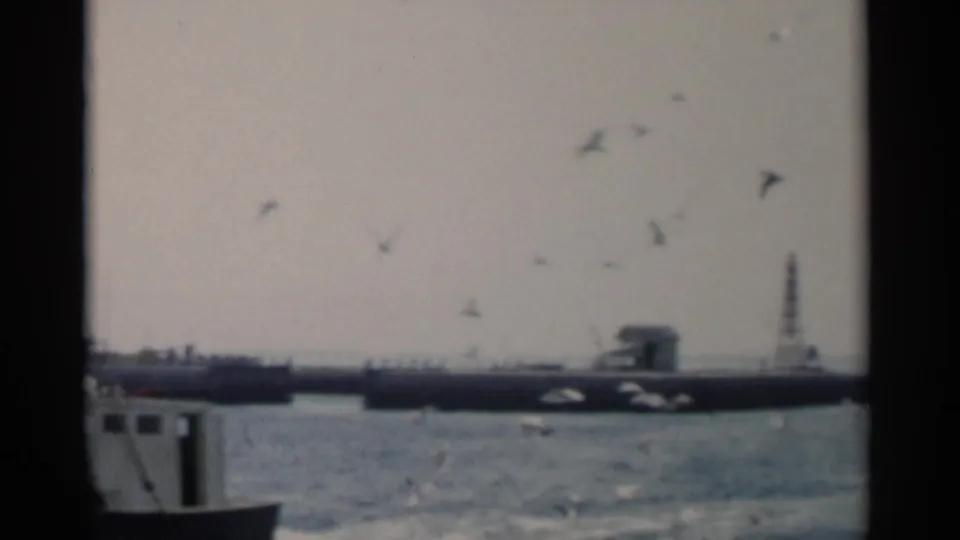 The camera catches part of a boat at frame edge while gulls fly over choppy Cape Cod water and a long pier or breakwater remains visible beyond.