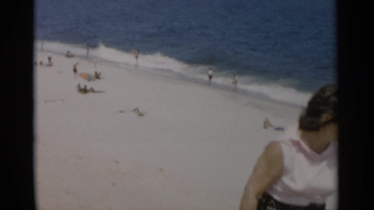 Beachgoers on white sand beside blue ocean surf with a woman partly visible at the right edge.