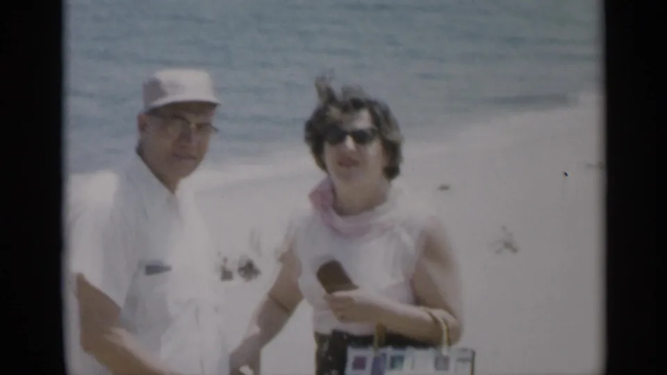 1956 Cape Cod beach close view of a man in white and a woman in sunglasses holding a handbag, with surf and beachgoers behind them.