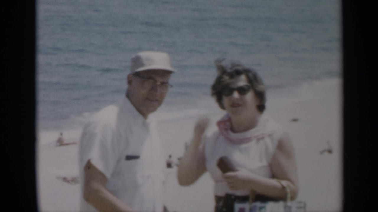 Man in white cap and woman in sunglasses stand on a sandy beach with ocean behind them.