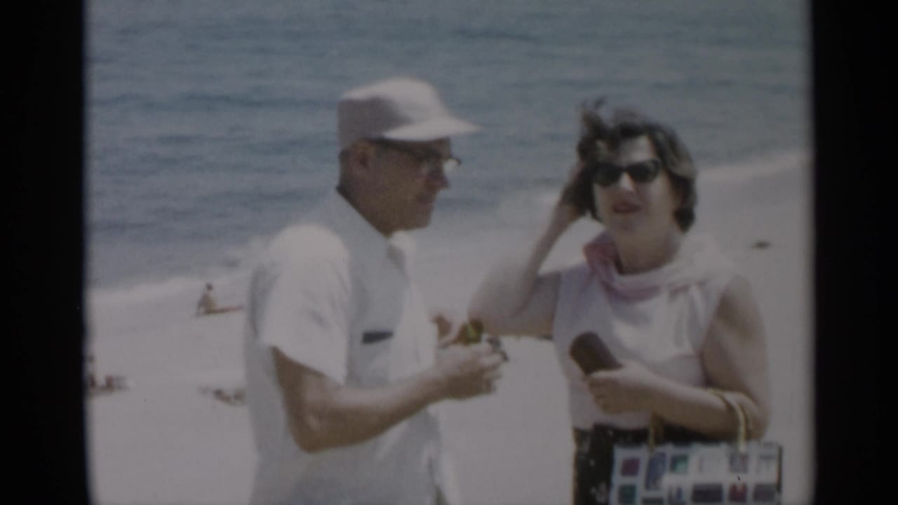 Man and woman stand near the surf as the woman lifts a hand to her hair.