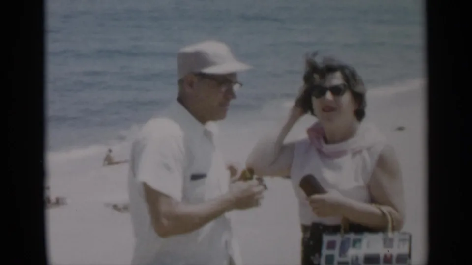 1956 Cape Cod beach frame with a man in white and a sunglassed woman adjusting her hair near the ocean.