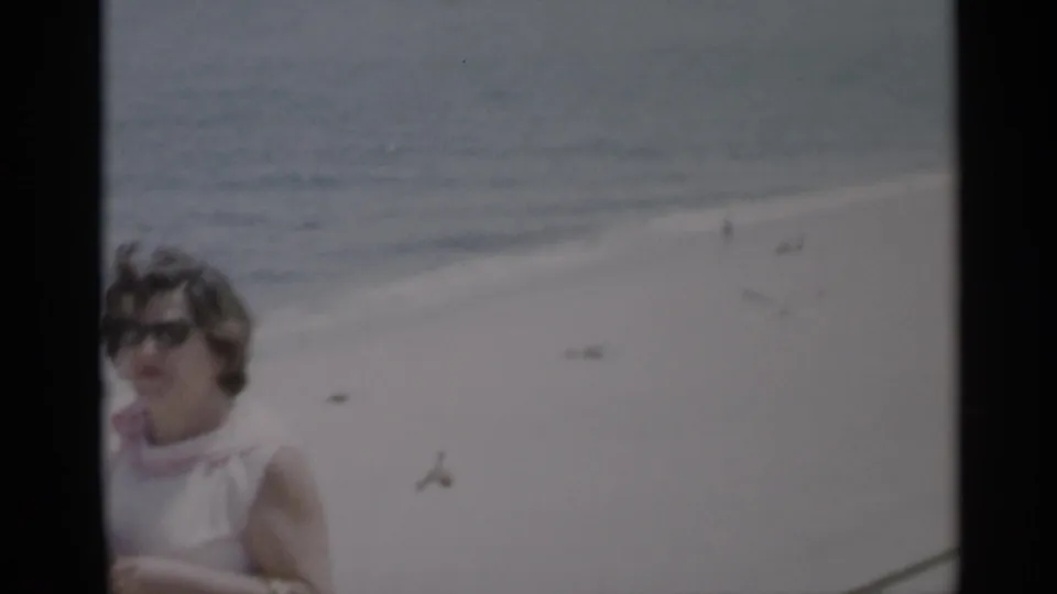 1956 Cape Cod beach frame with the woman partly cropped in front of open sand, distant figures, and blue water.
