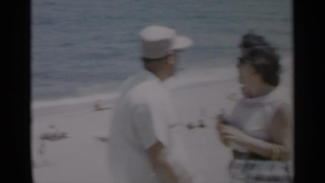 Man in a white cap faces a woman near a beach with ocean and bathers behind them.