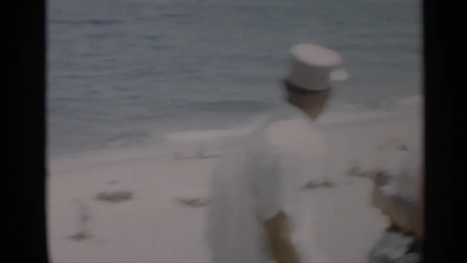The man in a white cap remains in the foreground of a soft 8mm Cape Cod beach scene, with bathers and surf behind him.