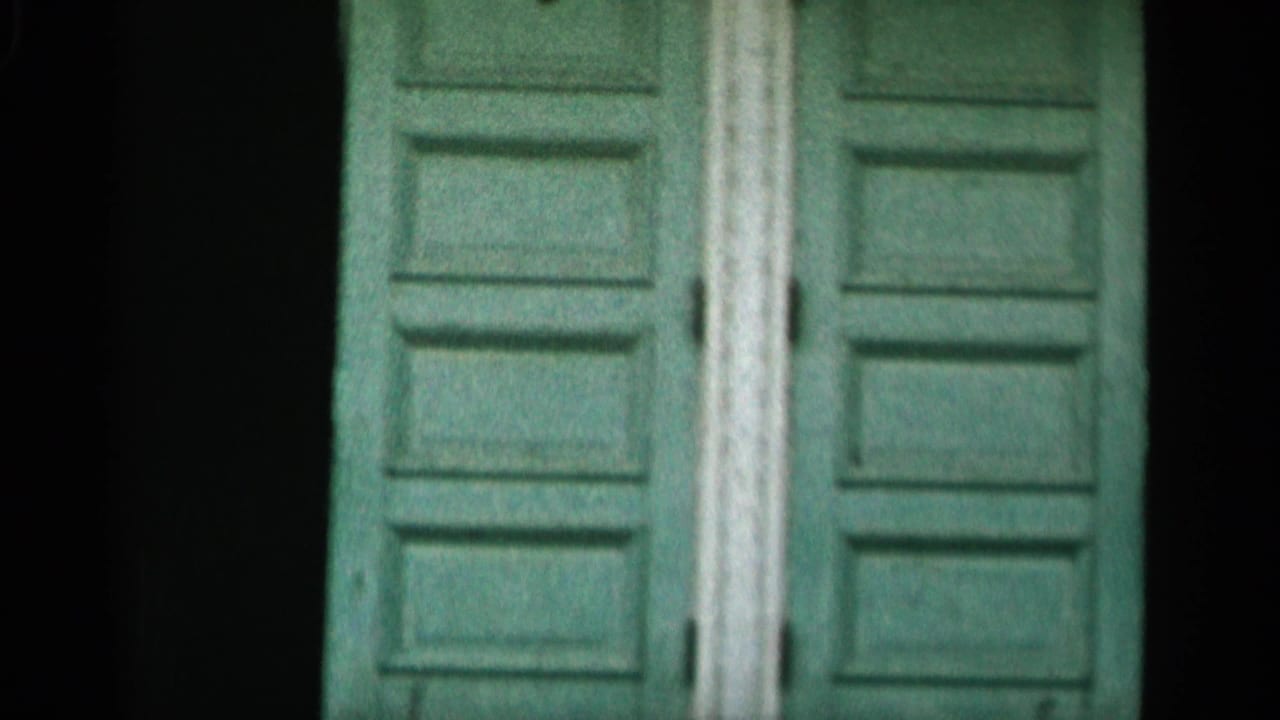 1956 Cape Cod 8mm frame showing weathered green double doors, pale vertical trim, and shadowed areas around the entrance.
