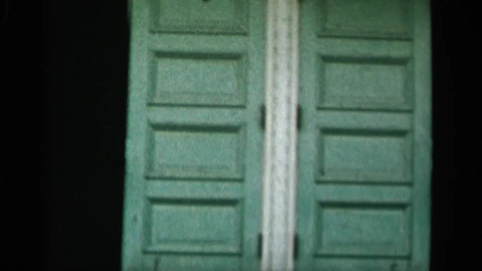 1956 Cape Cod 8mm frame showing weathered green double doors, pale vertical trim, and shadowed areas around the entrance.