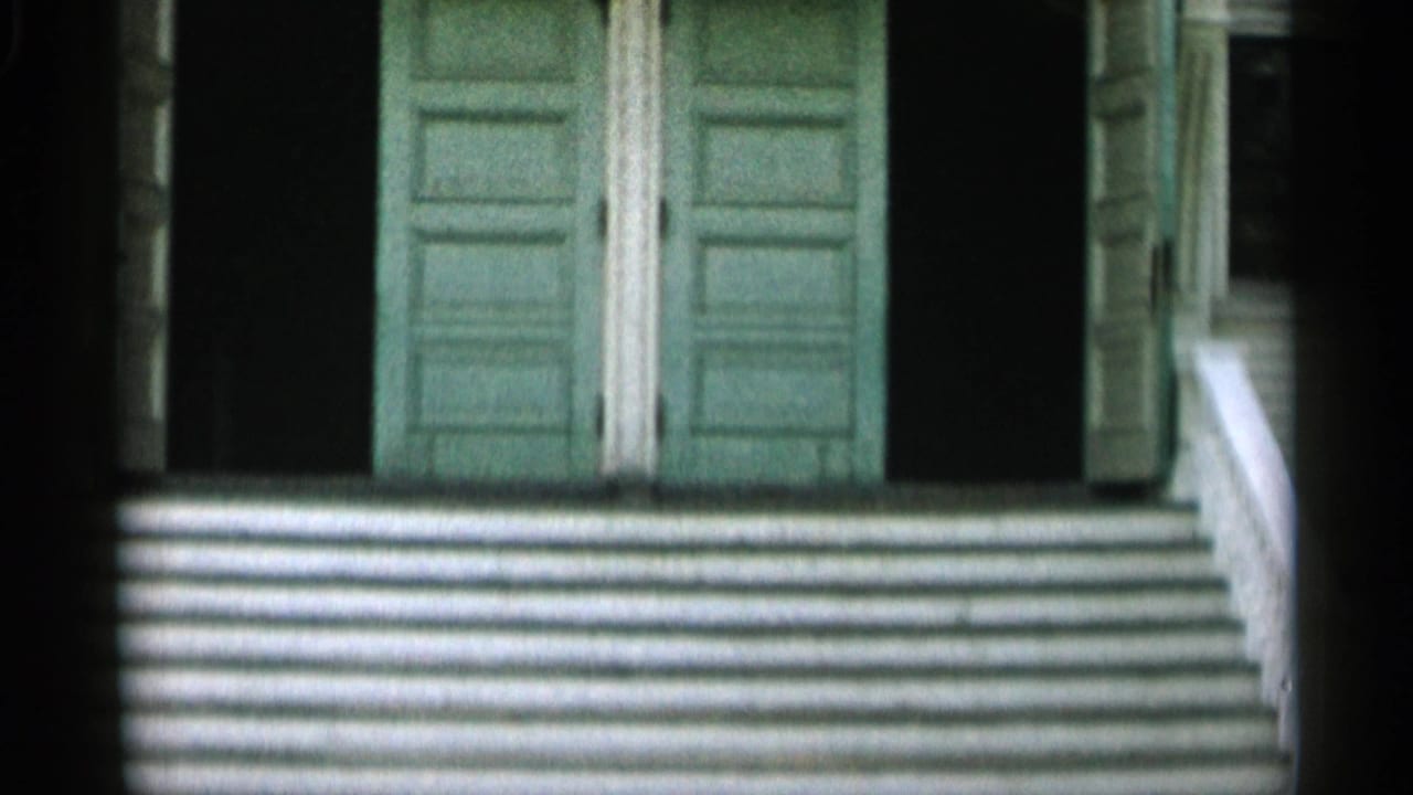 1956 Cape Cod home-movie frame with green doors at the top of white porch steps and deep shadows on both sides.