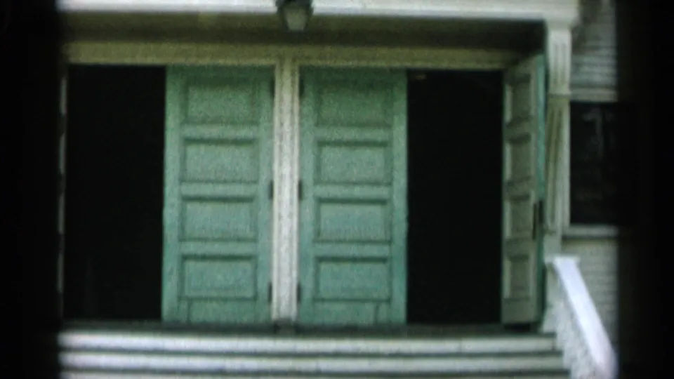 1956 Cape Cod frame showing a wider entrance view with green doors, clapboard siding, side shadows, white steps, and a partial railing.