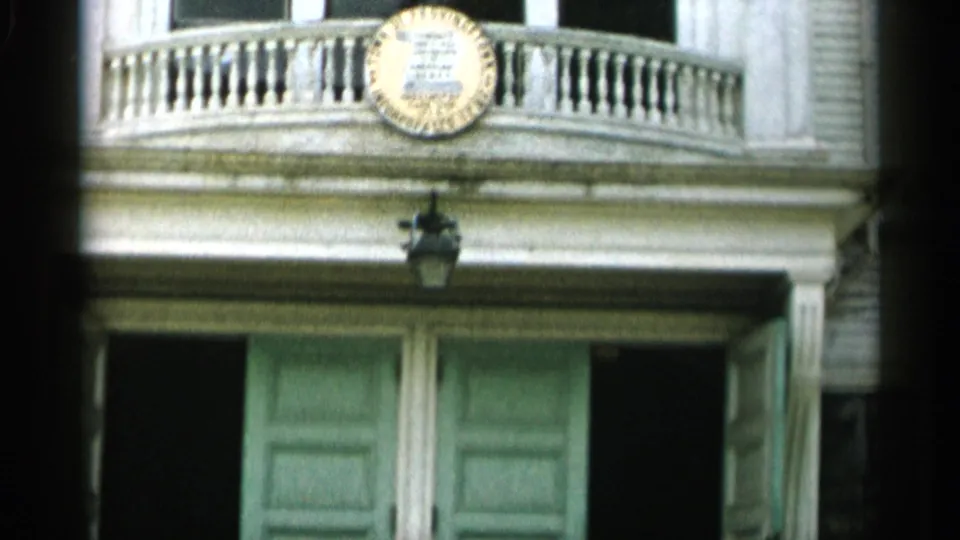 1956 Cape Cod 8mm frame showing the entrance facade with a balcony rail, circular emblem, hanging lantern, green doors, and white trim.