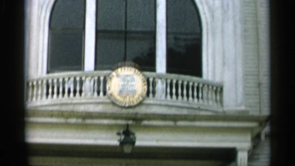 1956 Cape Cod home-movie frame showing a white facade with tall arched windows, balcony railing, and a circular plaque.