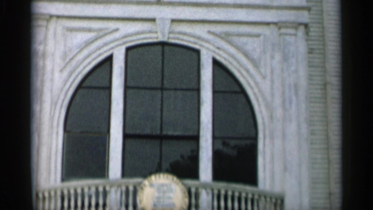 1956 Cape Cod frame with an arched upper window, white trim, balcony railing, and partially visible circular plaque.