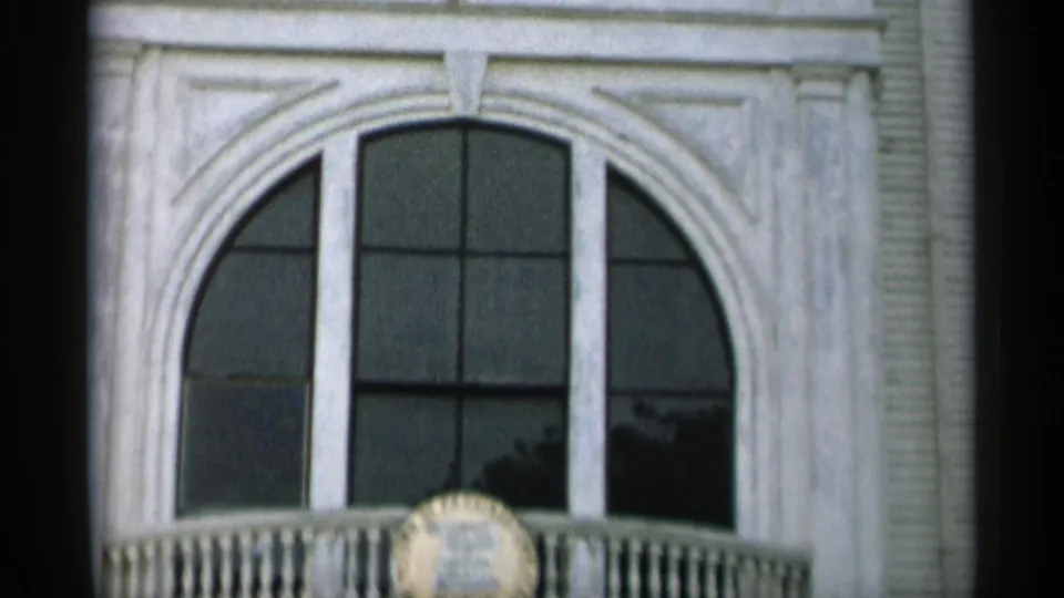 1956 Cape Cod frame with an arched upper window, white trim, balcony railing, and partially visible circular plaque.