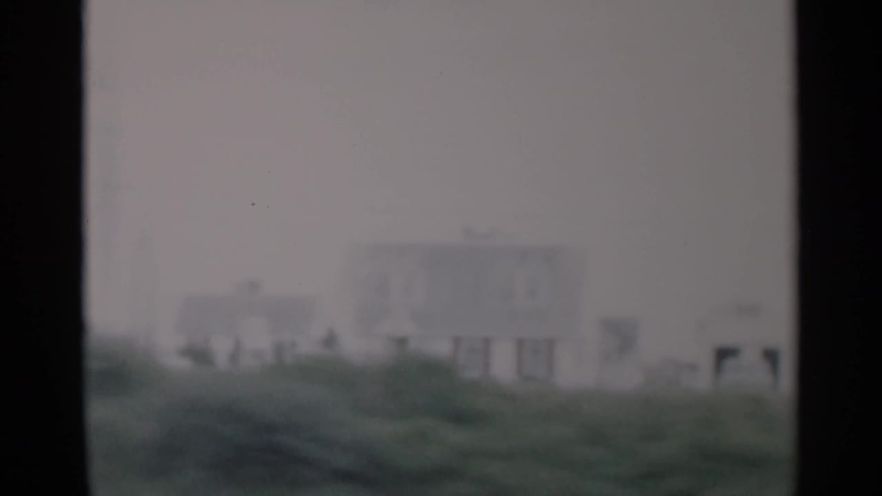 Soft, distant view of a pale house beyond green coastal grass under a cloudy sky.