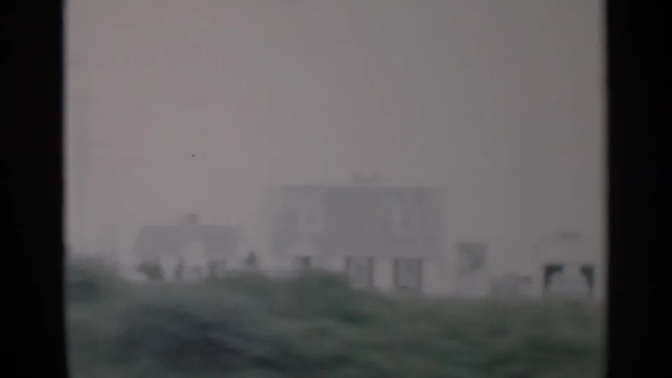 1956 Cape Cod 8mm frame showing a distant white house or building behind grassy dunes on an overcast day.