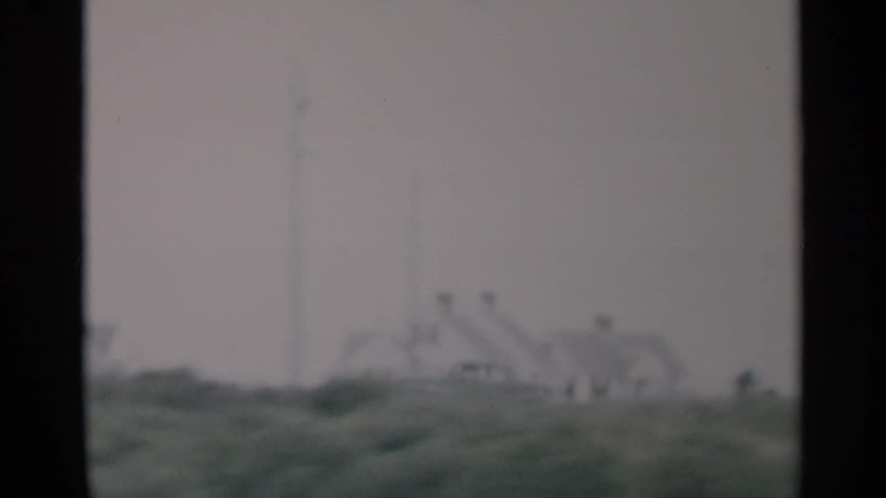 1956 Cape Cod frame with distant houses, low green dunes, and a pale overcast sky, softened by motion and 8mm focus.