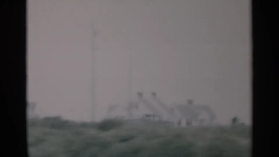 1956 Cape Cod frame with distant houses, low green dunes, and a pale overcast sky, softened by motion and 8mm focus.