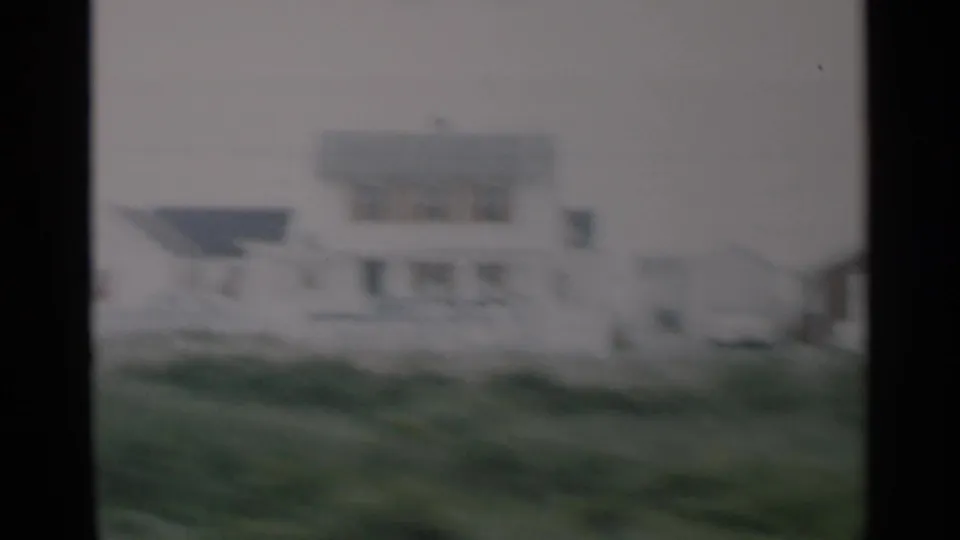 1956 Cape Cod 8mm frame showing a white residential building with a dark roof beyond blurred green foreground grass.