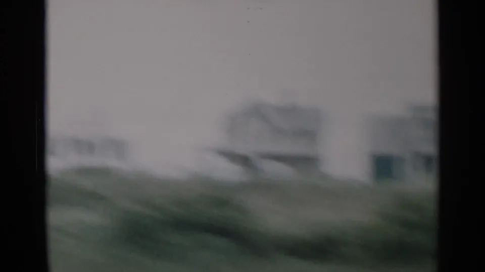 Soft, blurred 8mm frame of Cape Cod houses and roadside vegetation filmed from a moving vehicle.
